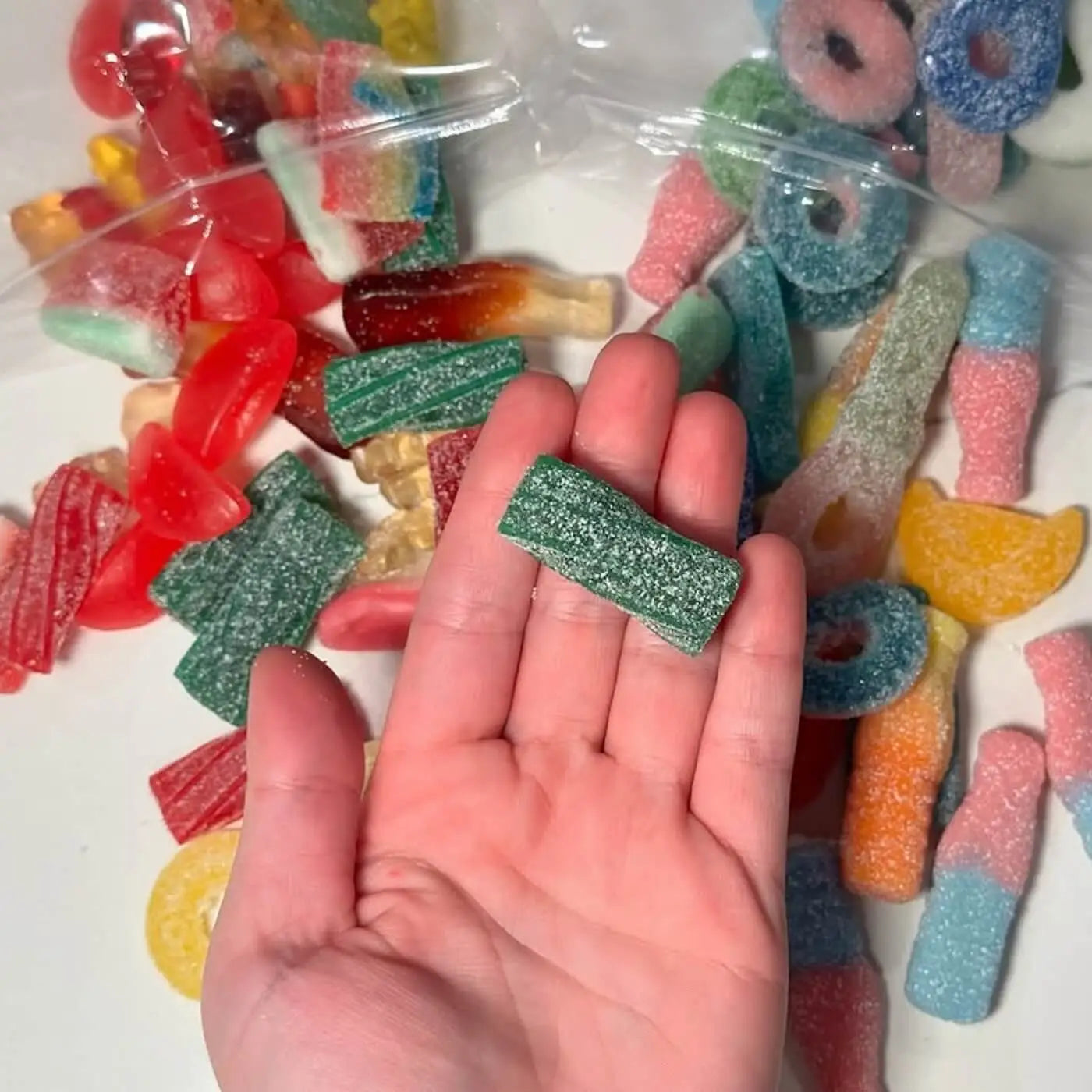 A piece of Swedish candy, green and coated in sugar, in a human hand, against a background of multiple pieces of candy on a table.